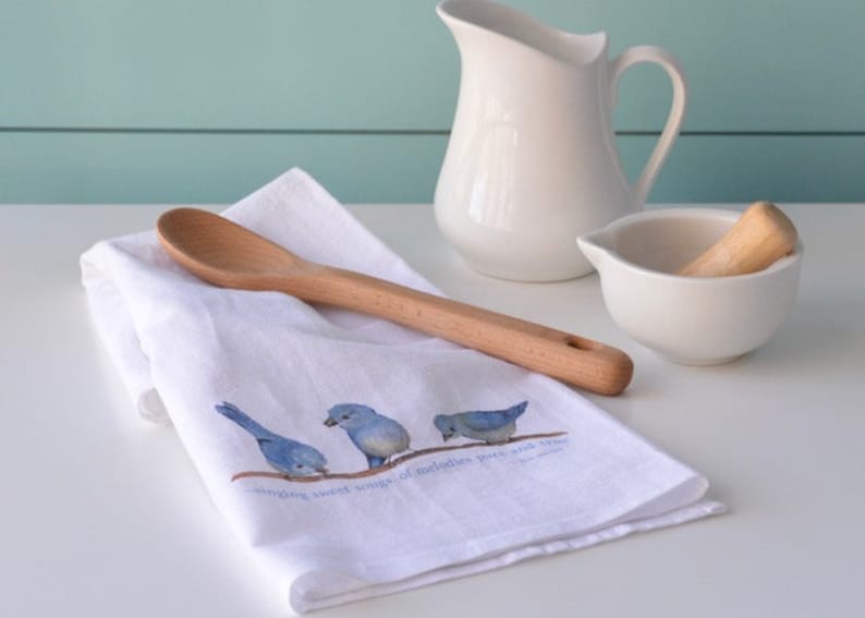 May include: White kitchen towel with a bluebird design and the text "singing sweet songs, of melodies pure and true". A wooden spoon is lying on top of the towel.