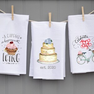 May include: Three white kitchen towels with different designs. The first towel features a cupcake with pink frosting and the text "A cupcake is happiness with icing on top". The second towel features a white wedding cake with pink flowers and the text "est. 2020". The third towel features a bicycle with pink balloons and the text "Celebrate the little things".