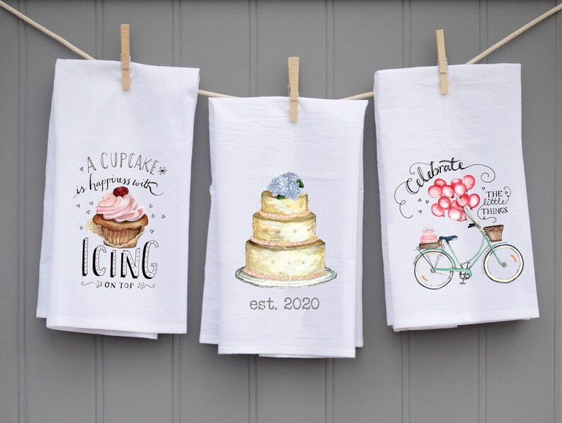 May include: Three white kitchen towels with different designs. The first towel features a cupcake with pink frosting and the text "A cupcake is happiness with icing on top". The second towel features a white wedding cake with pink flowers and the text "est. 2020". The third towel features a bicycle with pink balloons and the text "Celebrate the little things".