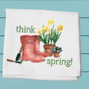 May include: White kitchen towel with a green and yellow illustration of a pair of red rain boots, a trowel, a hand rake, and a potted plant with yellow daffodils. The text "think spring!" is written in green.