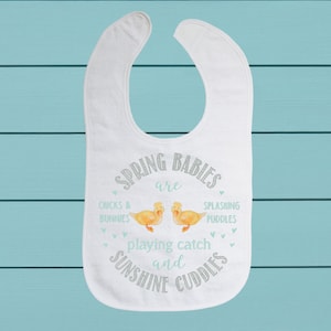 Puede incluir: Babero blanco para bebé con el texto "Spring babies are chicks & bunnies playing catch and sunshine cuddles" con dos patitos amarillos en el centro.