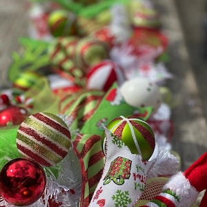 May include: A decorative garland featuring red, lime green, and white ornaments, including striped and solid spheres. The arrangement incorporates white tinsel, patterned ribbons with gift box and snowflake motifs, and a plush red and white hat element.