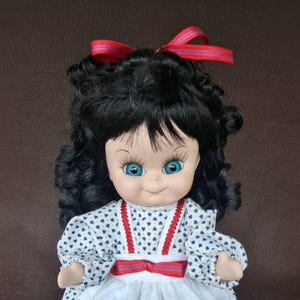 May include: A vintage doll with black curly hair and blue eyes. The doll wears a white dress with blue heart patterns and red trim. A red ribbon adorns the hair and a red bow accents the waist. The doll has red shoes.