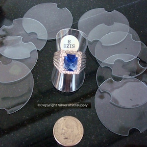 May include: Eight clear plastic ring display discs with a cut-out for a ring. The discs are stacked on top of each other. A ring with a large blue gemstone is displayed on a ring stand.