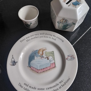 May include: A white ceramic set featuring Peter Rabbit illustrations, including a round plate with text about Peter going to bed, a hexagonal money box with a coin slot, and a small cup, all decorated with scenes of a rabbit and garden items.