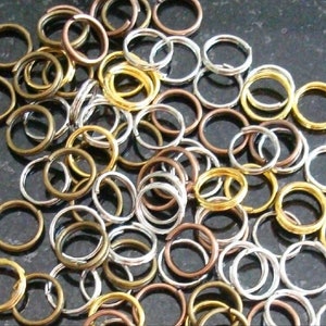 May include: A collection of split key rings in various metallic finishes, including gold, silver, and bronze. The rings are round and appear to be made of metal. They are scattered across a dark surface.