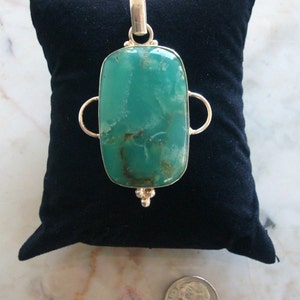 Puede incluir: Un colgante de plata con una piedra preciosa verde rectangular grande. El colgante tiene dos bucles a cada lado de la piedra.