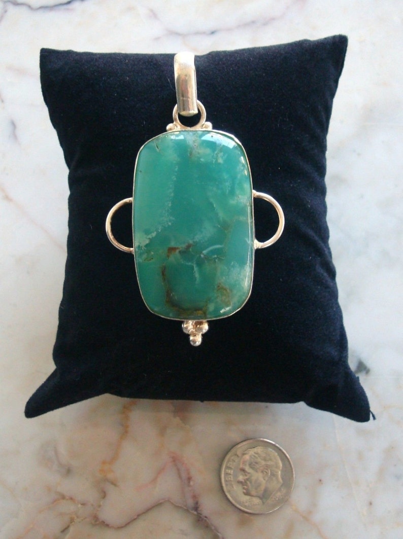 Puede incluir: Un colgante de plata con una piedra preciosa verde rectangular grande. El colgante tiene dos bucles a cada lado de la piedra.