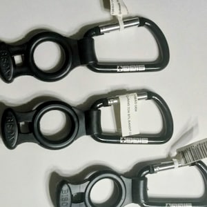 May include: Three black carabiner clips with a D-ring design. Each clip has a silver gate and the word "SIGGIE" printed on the side. The clips are connected to a black plastic piece with the word "BISON" printed on it.