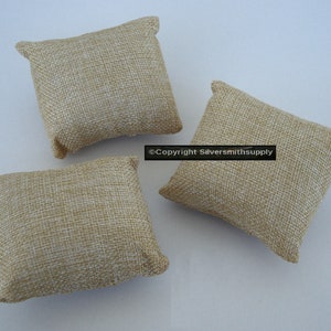 May include: Three small, square, beige burlap pillows with a textured weave. Each pillow has a slightly raised edge on each corner. The pillows are arranged on a white surface. The image is well-lit, highlighting the natural texture of the fabric.