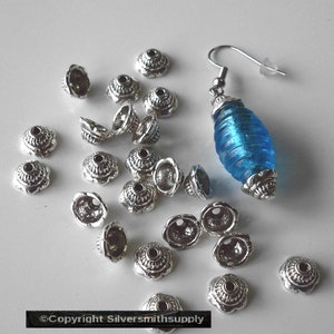May include: An earring with a blue glass bead and silver-colored metal accents. The earring is displayed with several silver-colored metal bead caps. The bead caps have a floral design.