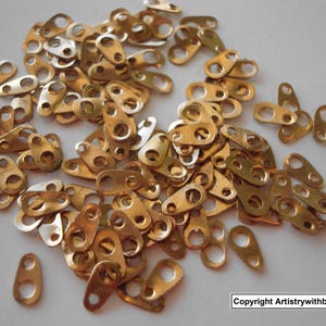 May include: A pile of small, gold-colored metal connectors with multiple holes. The connectors are in various shapes, including oval and rectangular, and are scattered across a light-colored surface. The image includes the text "Copyright Artistrywithbeads.shop".