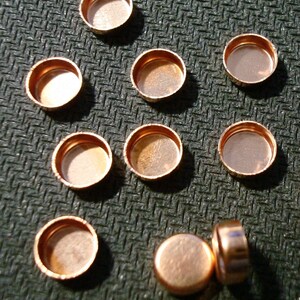 May include: A group of 10 copper-colored metal discs, some stacked on top of each other. The discs are round and have a smooth surface. They are arranged in a semi-circle on a dark gray textured surface.