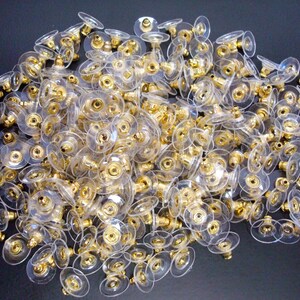 May include: A pile of clear earring backs with gold-colored metal posts and accents. The earring backs have a flat, circular design with a small post in the center. The gold accents add a touch of elegance to the otherwise simple design.