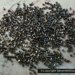 May include: A large pile of small, black metal jump rings. The rings are scattered on a white marble surface.