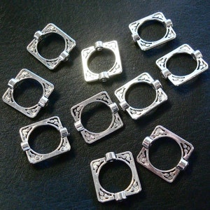 May include: Ten silver-toned metal square-shaped rings with ornate designs. Each ring has a circular opening in the center.