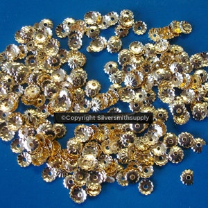 May include: A collection of small, gold-colored metal bead caps with a scalloped edge and a flower-like design. The bead caps are scattered across a blue surface, creating a textured appearance. The image is well-lit, highlighting the metallic sheen of the bead caps.