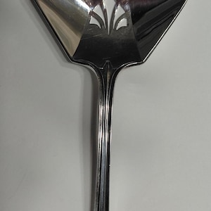 May include: A silver-colored serving utensil with a decorative, openwork design. The utensil has a wide, geometric head with a stylized floral pattern and a long, slender handle. The overall shape is symmetrical and elegant.