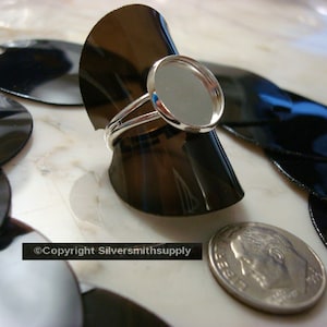 May include: A silver ring with a round, clear glass bezel setting. The ring is displayed on a black ring mandrel. The ring is surrounded by black, round, flat pieces of material.