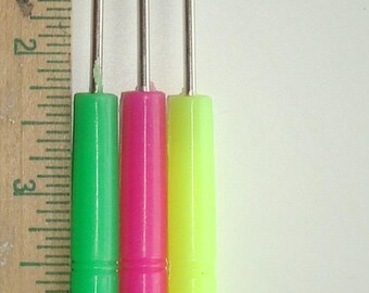 May include: Three beading needles with colorful handles in green, pink, and yellow. A wooden ruler is on the left side of the image. The needles are approximately 2 inches long.