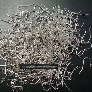 May include: A pile of silver-colored earring hooks, scattered on a dark surface. The hooks are made of thin wire and have a curved design for securing to the ear. The image shows a close-up view of the hooks.