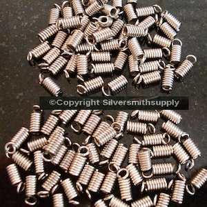 May include: A collection of silver-colored, coiled spring clasps with small loops at the ends. The clasps are tightly wound and appear to be made of metal. The image is a close-up shot, showing the details of the clasps. The clasps are likely used for jewelry making.