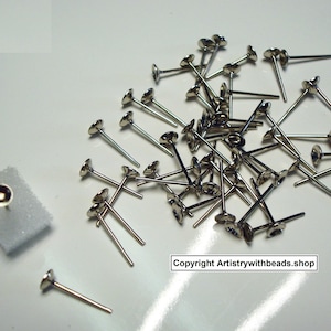 May include: A selection of silver-coloured metal earring posts with flat, round tops. The posts are scattered on a white surface, with one post attached to a small piece of white foam. The image includes the text "Copyright Artistrywithbeads.shop".