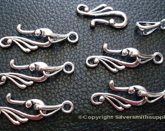 May include: A collection of silver-toned jewelry clasps in various sizes. The clasps feature an ornate, flowing design with curved elements and small circular details. Some clasps have a hook closure, while others have a loop.