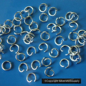 Puede incluir: Una gran cantidad de anillos de salto de plata, esparcidos sobre una superficie azul. Los anillos de salto son pequeños y circulares, con un espacio en el círculo. Se utilizan para la fabricación de joyas.