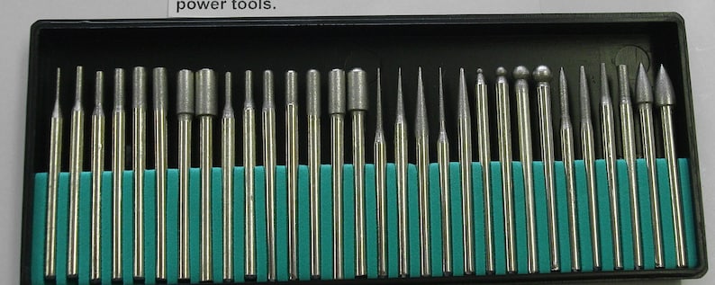 May include: A set of 25 silver diamond burrs in a black plastic case with a teal blue foam insert. The burrs are arranged in rows and are of various shapes and sizes.