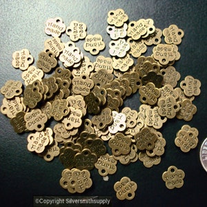 Op de afbeelding: Een stapel kleine, goudkleurige metalen labels met de woorden "Hand Made" erop gegraveerd. De labels zijn gevormd als een bloem met vier bloemblaadjes en hebben een gat in het midden om een ​​touwtje of lint aan te bevestigen.