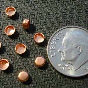 May include: A close-up of ten small, copper-colored, round, metal caps, arranged in a semi-circle pattern. A US dime coin is included for scale.