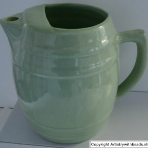May include: A light green ceramic pitcher with a handle and spout. The pitcher has a barrel-like design with horizontal ridges around the body. The handle is curved, and the spout is positioned at the top. The pitcher is likely used for serving beverages.