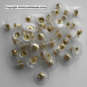 May include: A collection of clear earring backs with gold-colored posts and accents. The earring backs are made of a transparent plastic material and are scattered across a light-colored surface. The text "Copyright Artistrywithbeads.shop" is visible in the top left corner.