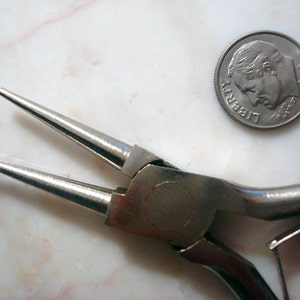 May include: Close-up of a pair of silver-toned jewellery pliers with red handles. The pliers are open and the jaws are visible. A US dime coin is in the background for scale.