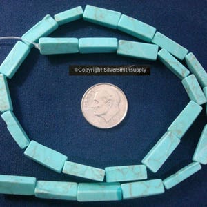 Peut inclure: Un brin de perles rectangulaires bleu turquoise, d'environ la taille d'une pièce de dix centimes d'euro, disposées en cercle sur une surface bleue. Le texte "Copyright Silversmithsupply" est visible sur l'image.