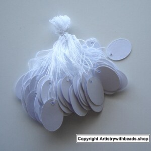 May include: A bundle of blank, white oval tags with a small hole at the top for stringing. The tags are attached to a white tassel. The background is a light gray surface. Copyright Artistrywithbeads.shop is visible.