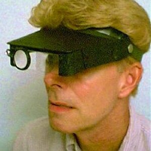 May include: A dark green visor with an attached magnifying lens and a small rectangular lens. The visor is worn on the head and has a strap. The person in the image has blond hair and is wearing a light-colored shirt.