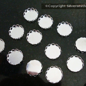 May include: A collection of silver-colored, scalloped bezel settings for jewelry making. The settings are round with a flat base and a decorative, wavy edge. The image is taken from above, showing the settings scattered on a dark surface.
