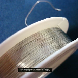 May include: A spool of silver wire on a white plastic bobbin. The wire is wound tightly and is ready to be used for jewelry making or other crafts.