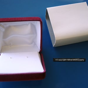 Könnte beinhalten: Eine Schmuckschatulle aus burgunderfarbenem Samt mit einem weißen Satin-Innenfutter. Die Schachtel ist geöffnet und hat zwei kleine Löcher im Boden für Ohrringe. Die Schachtel steht auf einer blauen Oberfläche. ©Copyright Silversmithsupply
