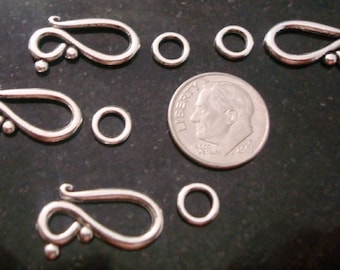 May include: Silver-toned jewelry clasps and jump rings are displayed. The clasps have a decorative swirl design with small ball accents. Several jump rings of varying sizes are also present. A U.S. dime is included for size reference.