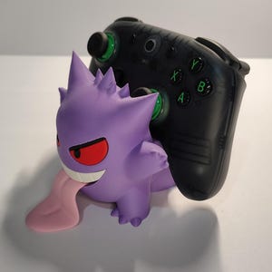 May include: A purple Gengar figure with red eyes and a pink tongue is positioned in front of a black video game controller. The controller has green accents on the buttons. The items are on a white surface.