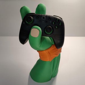 May include: A green hand-shaped gaming controller holder with an orange wristband, holding a black gaming controller with green accents. The holder is a unique and decorative item for gamers.