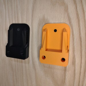 May include: Two plastic power tool holders, one black and one orange, mounted on a light wood surface. The holders have a rectangular shape with a slot for the tool and mounting holes.