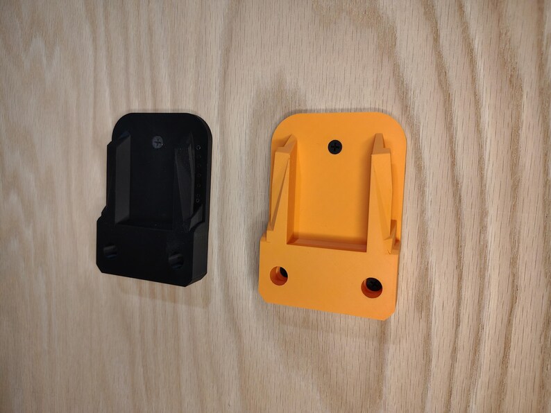 May include: Two plastic power tool holders, one black and one orange, mounted on a light wood surface. The holders have a rectangular shape with a slot for the tool and mounting holes.