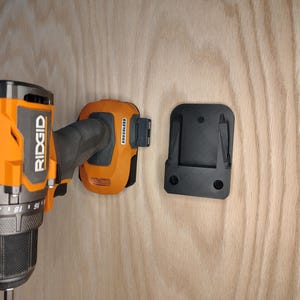 May include: An orange and black Ridgid cordless drill with a black battery and a black mounting bracket. The drill is mounted on a light wood surface. The battery is labeled "Brushless".