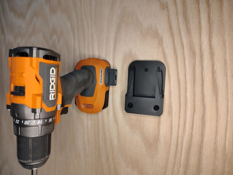May include: An orange and black Ridgid cordless drill with a black battery and a black mounting bracket. The drill is mounted on a light wood surface. The battery is labeled "Brushless".