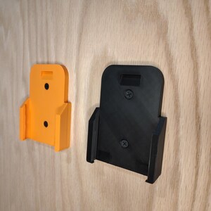 Puede incluir: Dos soportes de pared para herramientas el&eacute;ctricas. Uno es de color naranja brillante, el otro es negro. Ambos son de pl&aacute;stico y tienen forma rectangular con una ranura en la parte superior y una base curvada. Est&aacute;n montados sobre una superficie de madera clara.