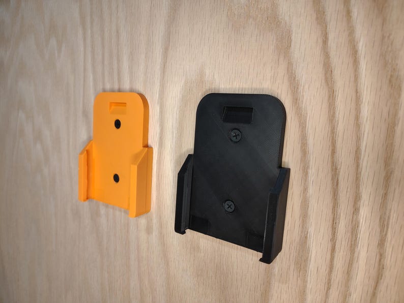 Puede incluir: Dos soportes de pared para herramientas el&eacute;ctricas. Uno es de color naranja brillante, el otro es negro. Ambos son de pl&aacute;stico y tienen forma rectangular con una ranura en la parte superior y una base curvada. Est&aacute;n montados sobre una superficie de madera clara.